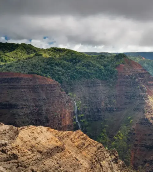 Red and green Waimea Canyon cliffs with a narrow waterfall under cloudy sky
