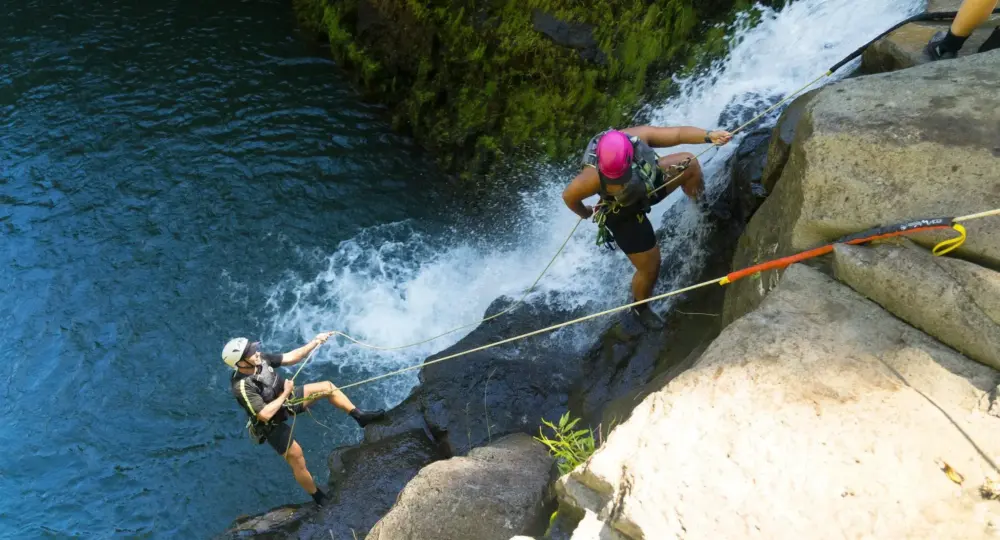 two people rappelling down a waterfall near mossy rocks over blue water
