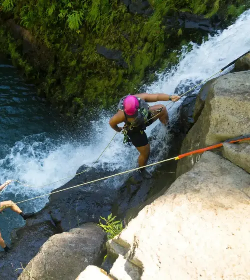 two people rappelling down a waterfall near mossy rocks over blue water