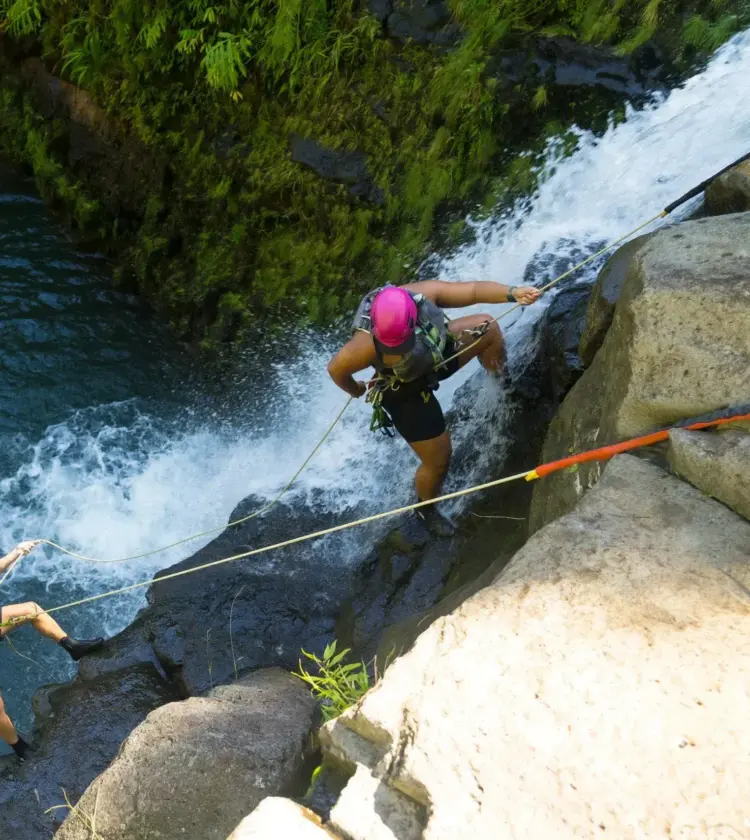 two people rappelling down a waterfall near mossy rocks over blue water