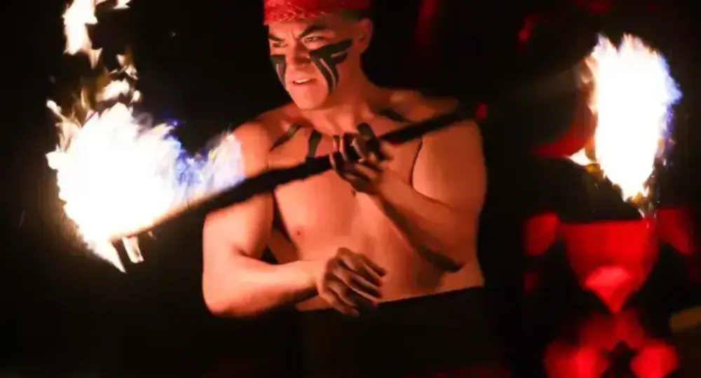 Bare-chested fire dancer wearing red headband and traditional skirt performing with flaming torches at night