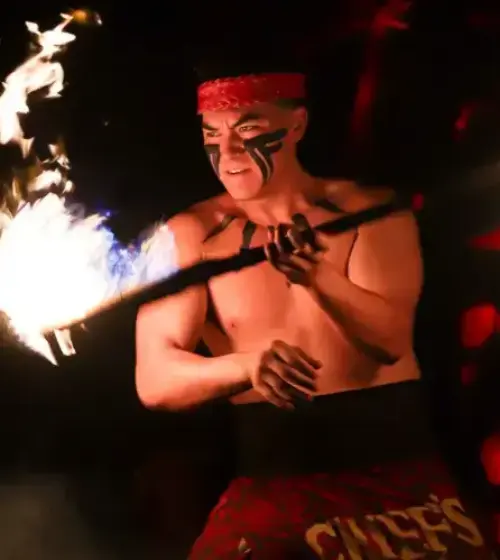 Bare-chested fire dancer wearing red headband and traditional skirt performing with flaming torches at night