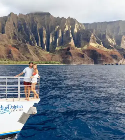 couple standing on boat deck with ocean and dramatic mountain cliffs in background