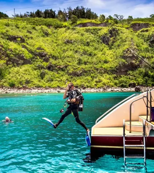 Scuba diver jumping off a boat into turquoise ocean with green cliffs in background