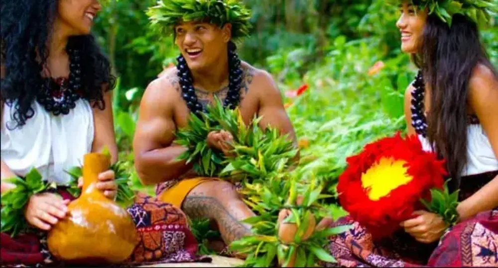 Three Hawaiians in traditional dress smiling and holding cultural items in a green forest setting