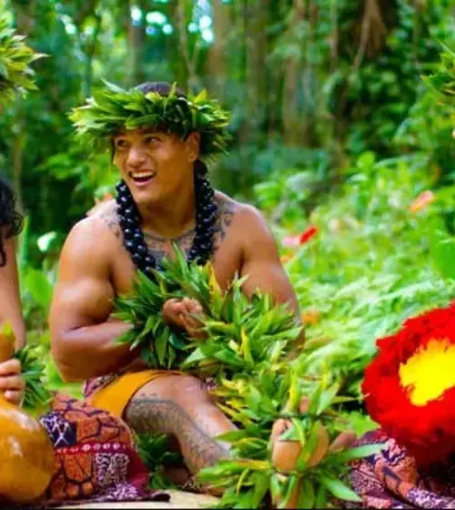 Three Hawaiians in traditional dress smiling and holding cultural items in a green forest setting