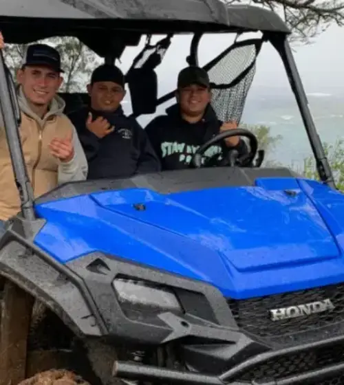 Three people seated in a blue Honda off-road vehicle on muddy ground near trees and ocean shore