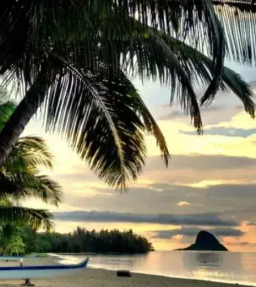 Hawaiian beach at sunset with palm trees, canoe on shore, and island silhouette in distance
