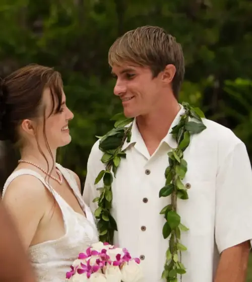 Smiling bride and groom looking at each other during Hawaiian wedding ceremony with person blowing conch shell in foreground