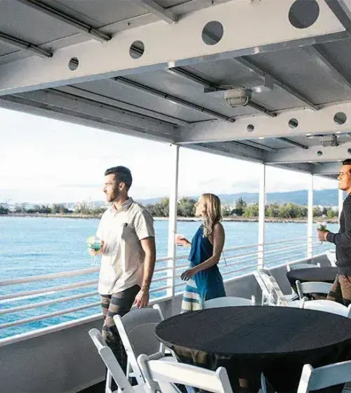 three people on a boat deck holding drinks with ocean and distant shoreline visible