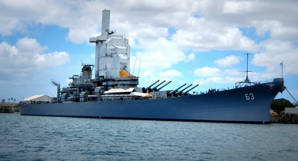 large blue battleship docked in calm harbor water under partly cloudy sky