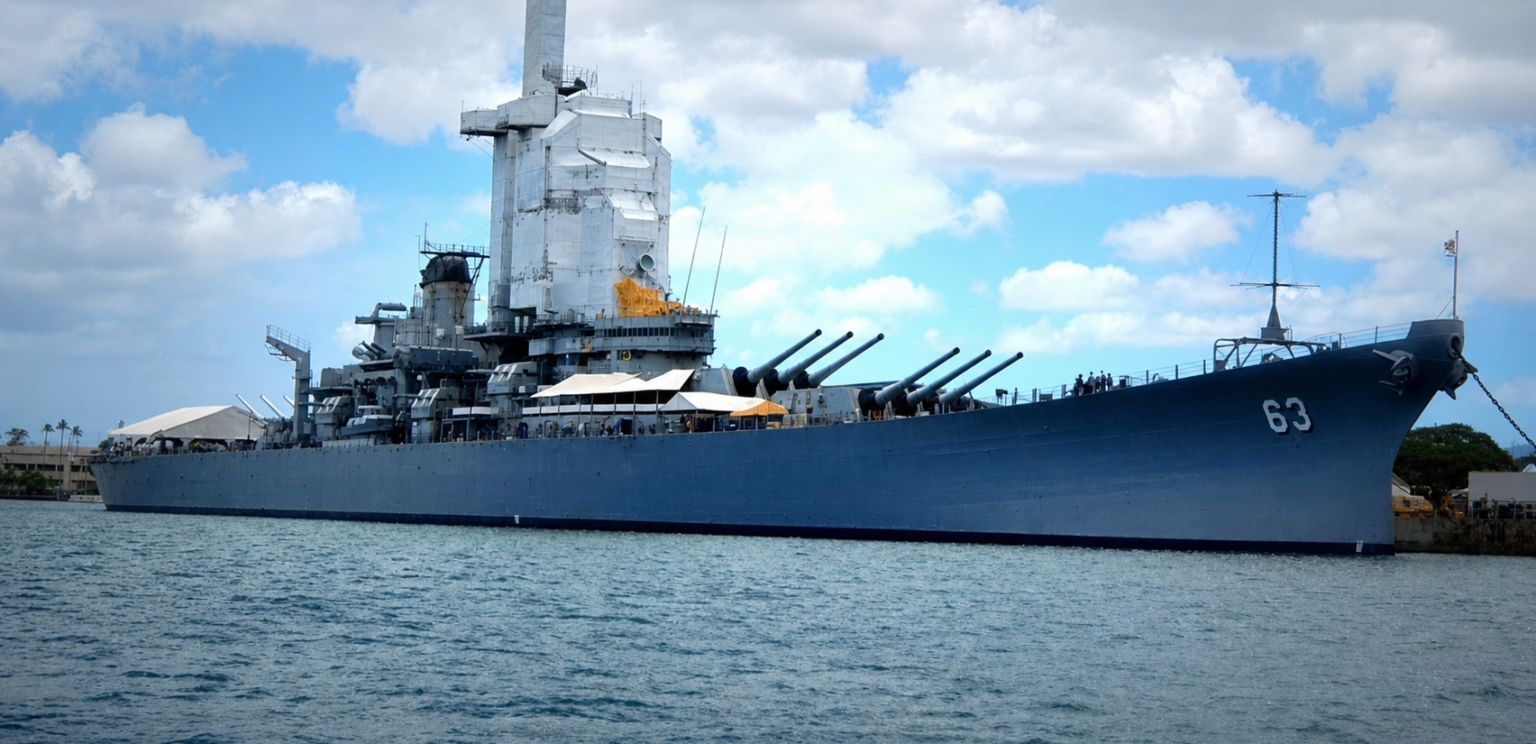 large blue battleship docked in calm harbor water under partly cloudy sky