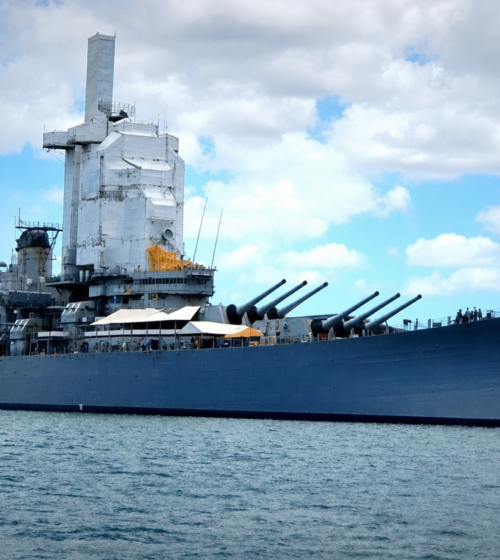 large blue battleship docked in calm harbor water under partly cloudy sky