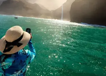 person wearing sun hat and blue floral shirt photographing Na Pali Coast cliffs with bright sun and turquoise sea