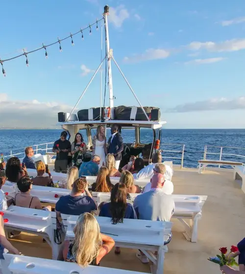 wedding ceremony with bride, groom, and guests seated on benches on a boat at sea under clear blue sky