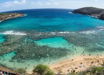 Clear turquoise bay with coral reef and sandy beach with people under a partly cloudy blue sky