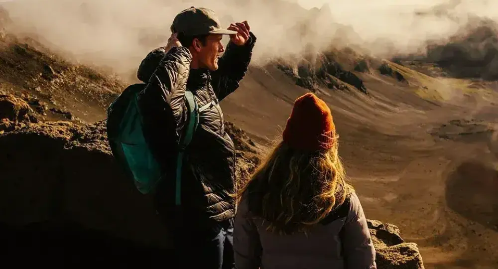 two hikers overlooking volcanic crater with mist and rocky terrain