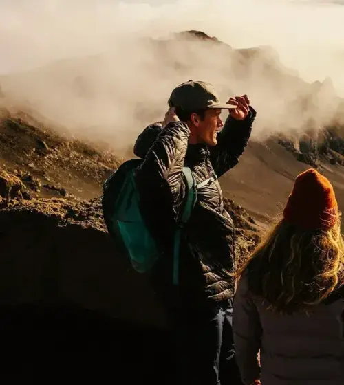 two hikers overlooking volcanic crater with mist and rocky terrain