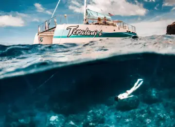 Catamaran named Teralani 4 on ocean surface with snorkeler swimming underwater over coral reef