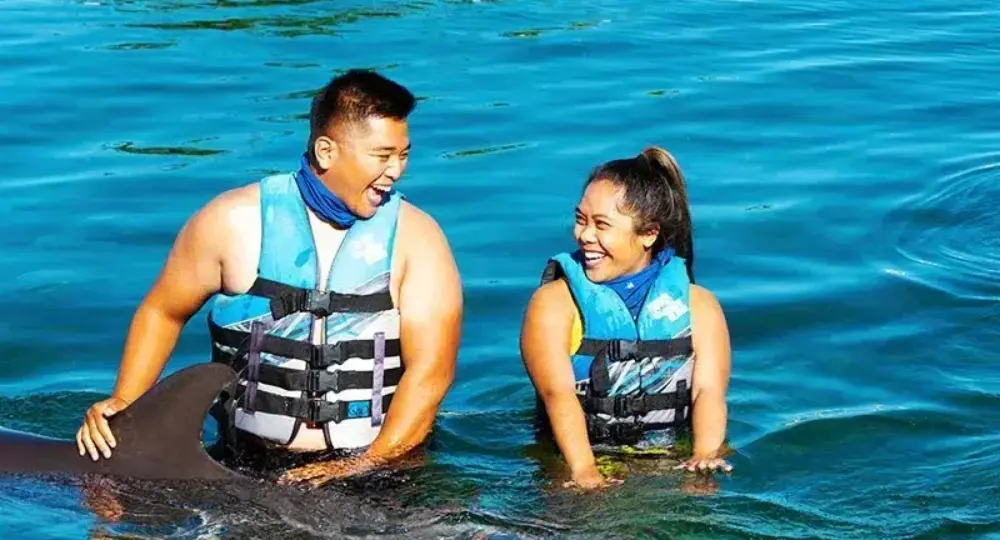 Smiling couple wearing life vests in blue water interacting with dolphin