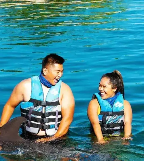 Smiling couple wearing life vests in blue water interacting with dolphin