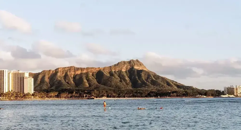 Diamond Head volcano in Honolulu with ocean and paddleboarders in foreground under cloudy sky
