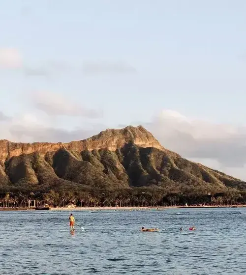 Diamond Head volcano in Honolulu with ocean and paddleboarders in foreground under cloudy sky