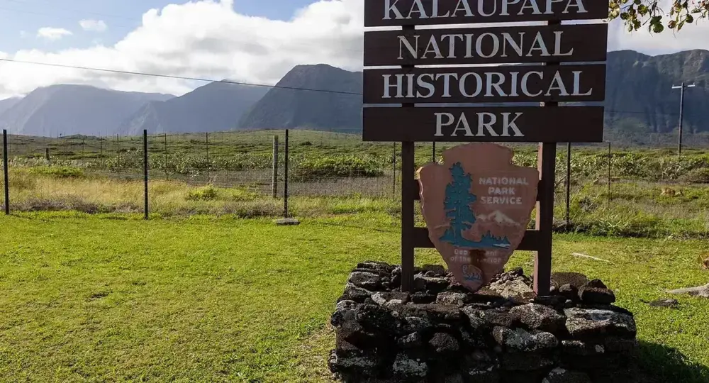 Kalaupapa National Historical Park wooden sign on stone base with green field and mountain range in the background