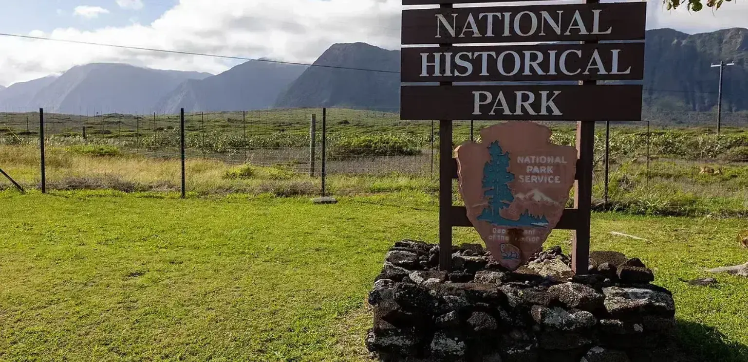 Kalaupapa National Historical Park wooden sign on stone base with green field and mountain range in the background