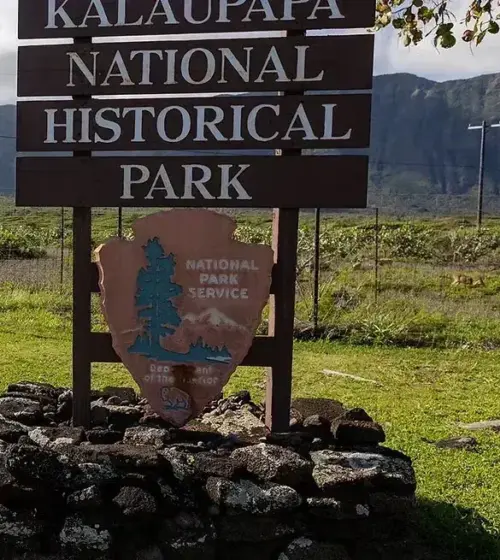 Kalaupapa National Historical Park wooden sign on stone base with green field and mountain range in the background