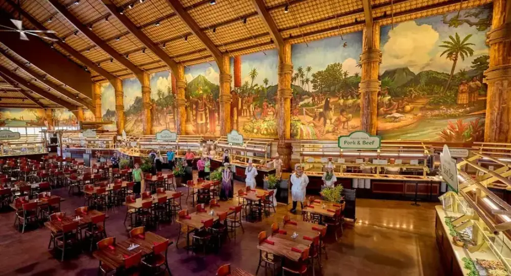 Spacious indoor dining area with wooden tables and chairs under a high ceiling featuring a large mural depicting Hawaiian scenes