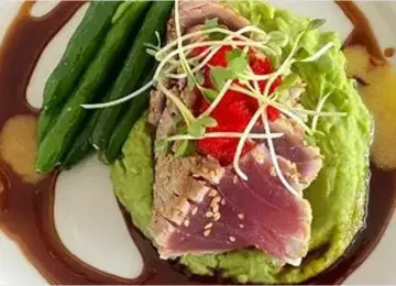 Seared tuna on avocado puree with green beans and garnished with sprouts and sauce on a white plate