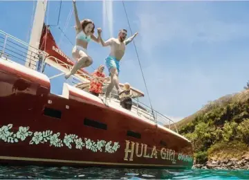 Two people jumping from a red boat named Hula Girl into clear ocean water with hillside in the background