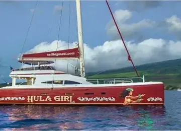 Red sailing catamaran named Hula Girl sailing on ocean near green hills under blue sky