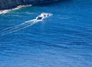 white catamaran boat sailing near steep rocky cliffs in blue ocean water