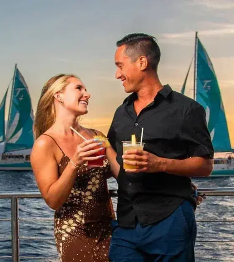 couple holding tropical drinks on sailboat at ocean sunset with two turquoise sailboats in background