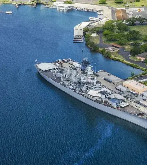 A naval battleship docked at a pier with surrounding harbor buildings and greenery
