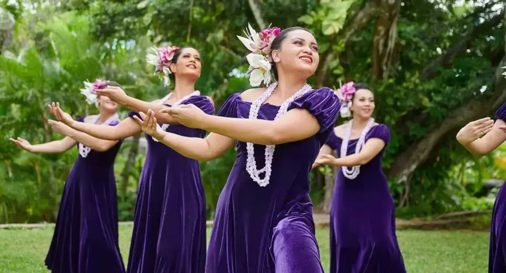 Hawaiian hula dancers wearing purple dresses, floral headpieces, and white leis performing outdoors with green trees in background