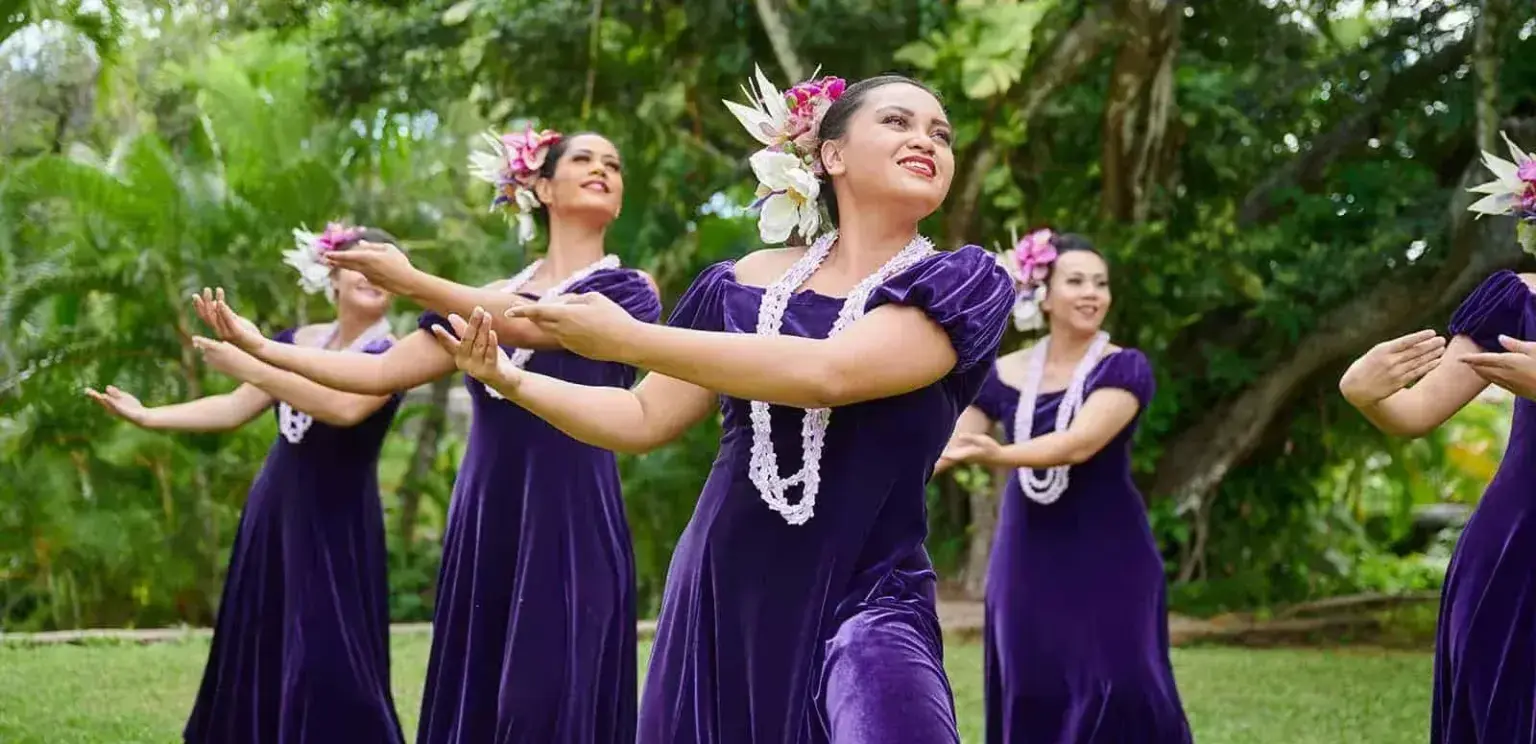 Hawaiian hula dancers wearing purple dresses, floral headpieces, and white leis performing outdoors with green trees in background