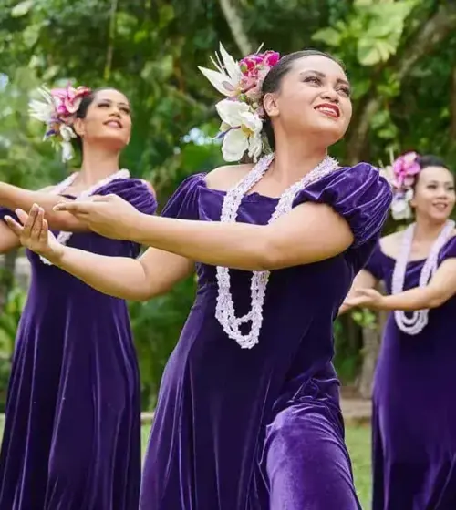 Hawaiian hula dancers wearing purple dresses, floral headpieces, and white leis performing outdoors with green trees in background