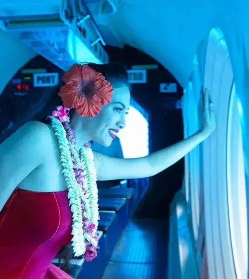 Woman wearing a red dress and lei with a large red flower in her hair looking out a submarine window