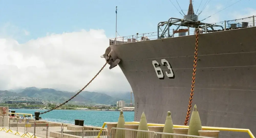 Large gray naval ship docked with visible hull number 63, chain anchors, harbor, and distant mountains under blue sky with clouds