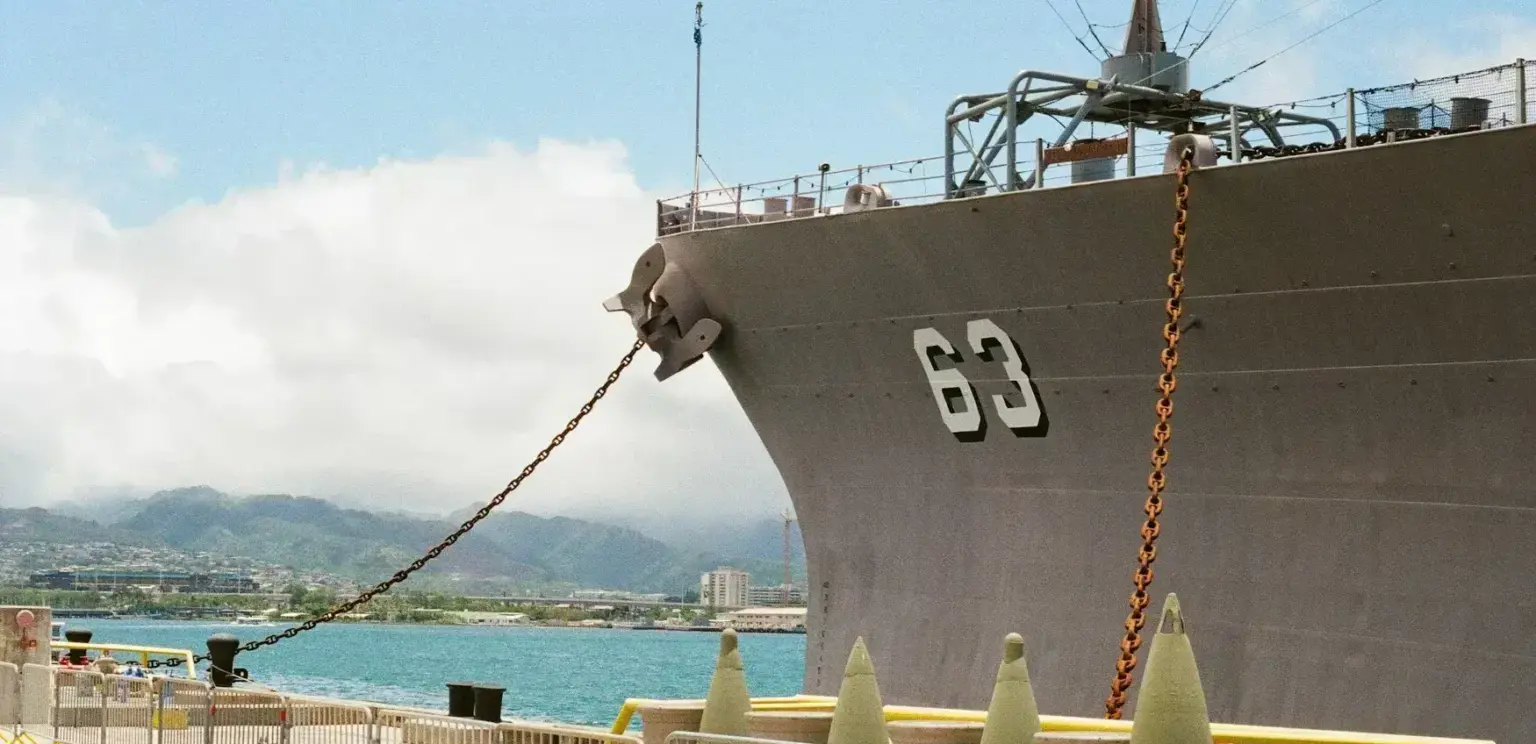 Large gray naval ship docked with visible hull number 63, chain anchors, harbor, and distant mountains under blue sky with clouds