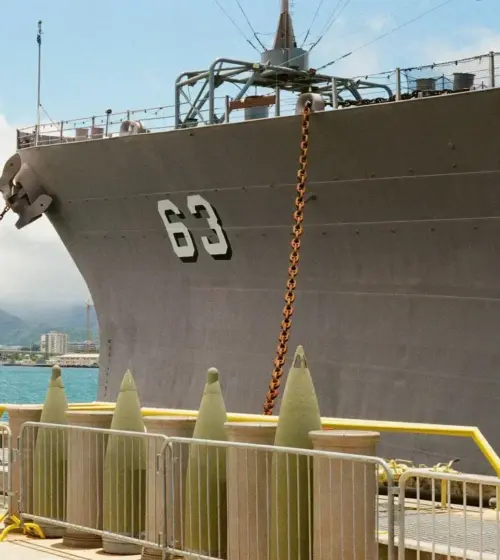 Large gray naval ship docked with visible hull number 63, chain anchors, harbor, and distant mountains under blue sky with clouds
