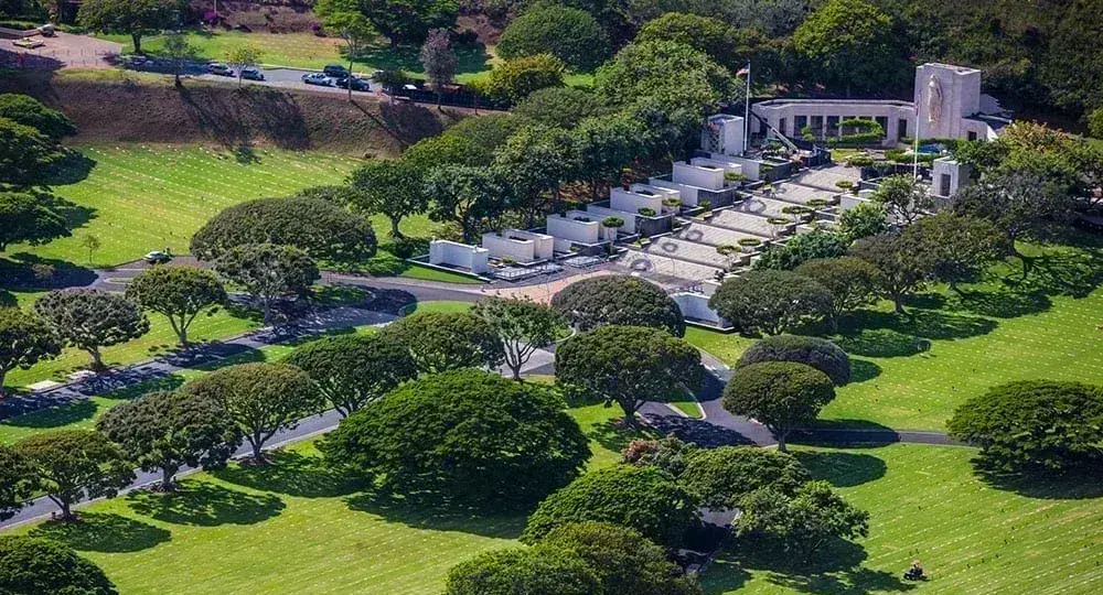 Aerial view of a memorial park with manicured green lawns, evenly spaced trees, pathways, and a white memorial structure.