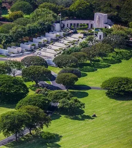 Aerial view of a memorial park with manicured green lawns, evenly spaced trees, pathways, and a white memorial structure.