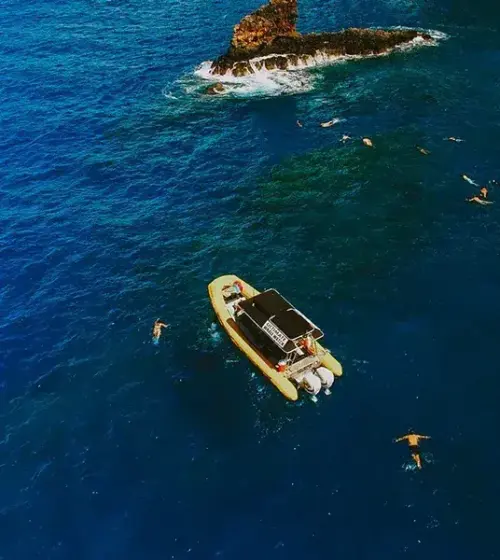 yellow boat floating in blue ocean with snorkelers swimming around near rocky outcrop