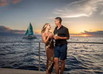 couple holding cocktails and enjoying sunset on a boat with sailboat and ocean in background