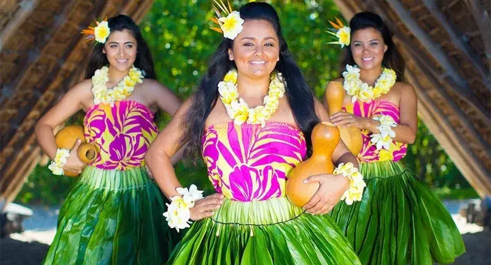 Three hula dancers in green grass skirts and pink floral tops holding traditional gourds