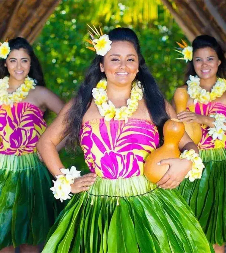 Three hula dancers in green grass skirts and pink floral tops holding traditional gourds